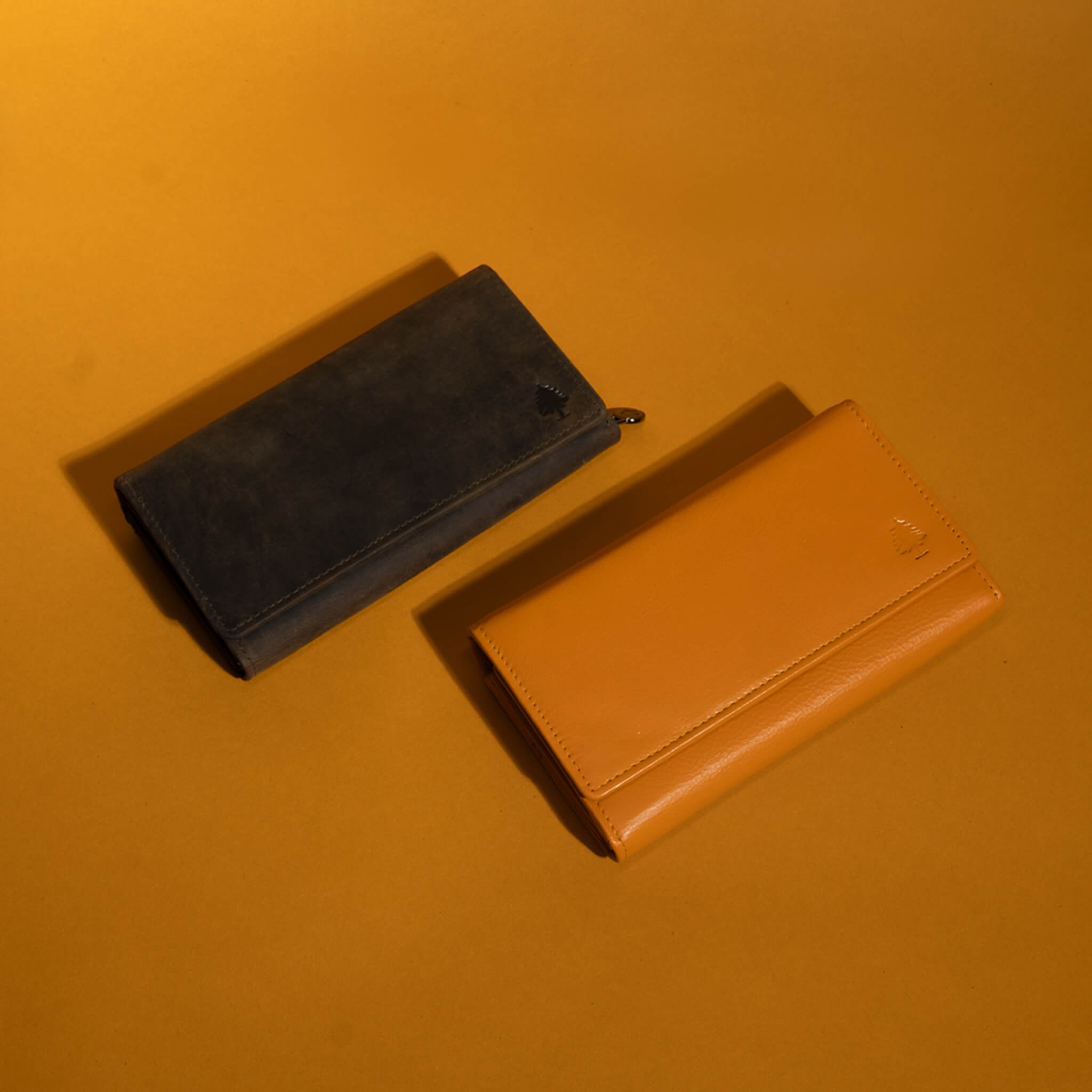 Zwei lange Leder-Geldbörsen auf warmgelbem Hintergrund – eine olivgrüne Wildleder-Geldbörse und eine glatte senfgelbe Leder-Geldbörse im minimalistischen Design